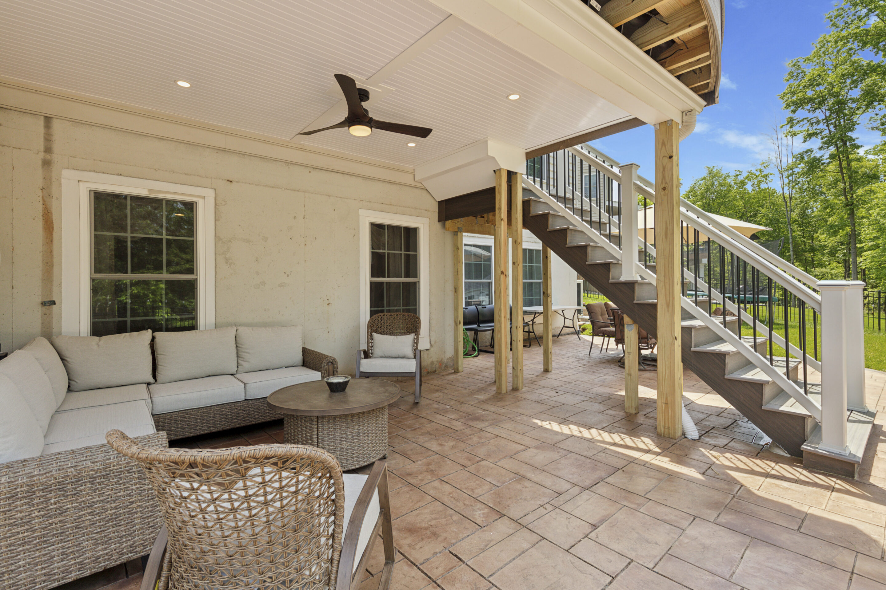 A custom-built deck in Warrington, PA with Trex composite decking and Trex Select railings, featuring a curved front edge. Below the deck, there is a waterproof lounge area with wicker furniture, a ceiling fan, and recessed lighting, creating a comfortable shaded retreat. The staircase leads to an outdoor seating area surrounded by greenery 40.2503° N, 75.1662° W