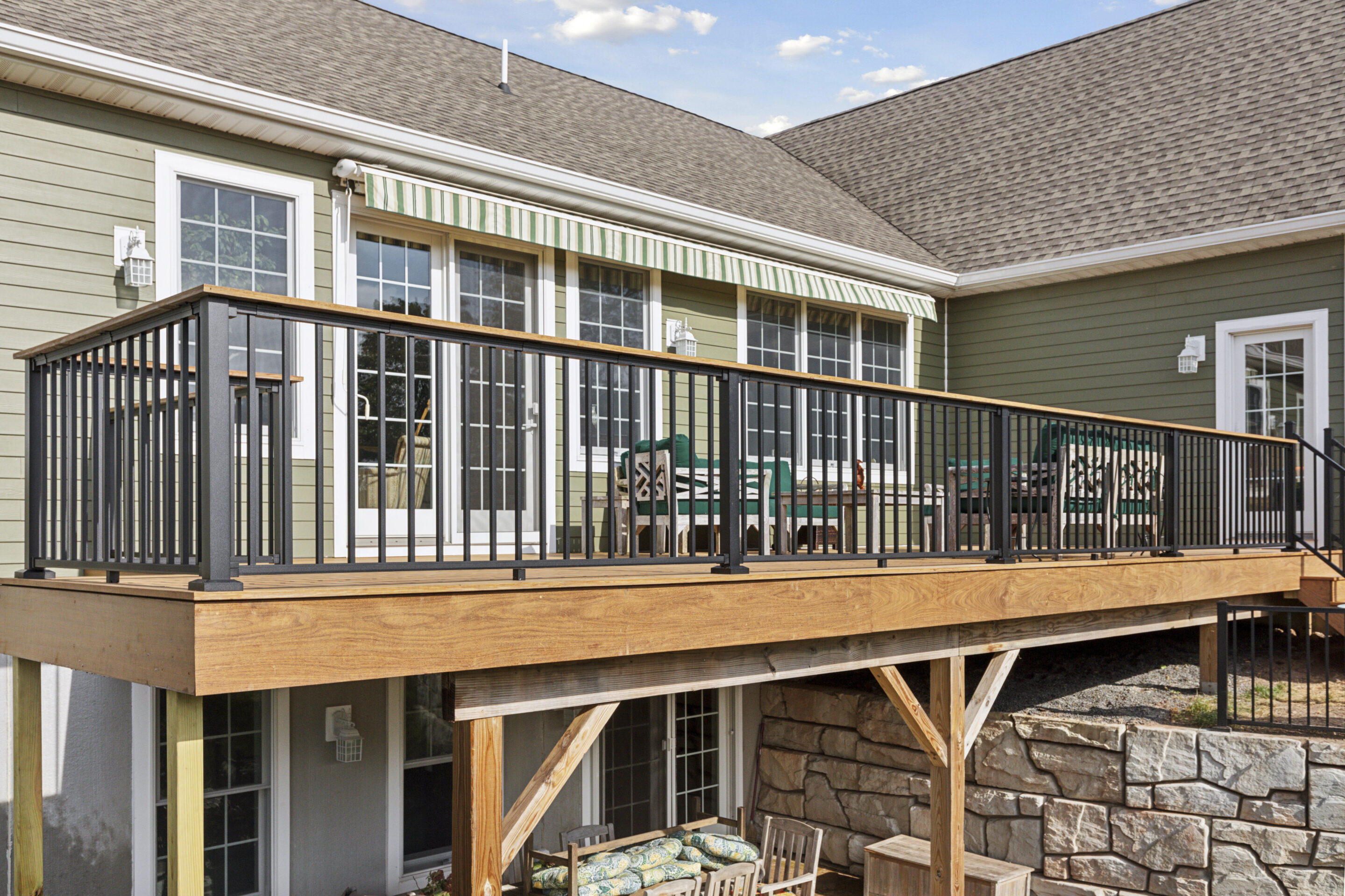 A custom-built elevated deck constructed with Brazilian Ipe lumber and black aluminum railings, featuring a green house with large windows and a striped awning in the background, and a stone retaining wall with a seating area underneath. Solebury, PA 18963 (40.4732° N, 75.1610° W)