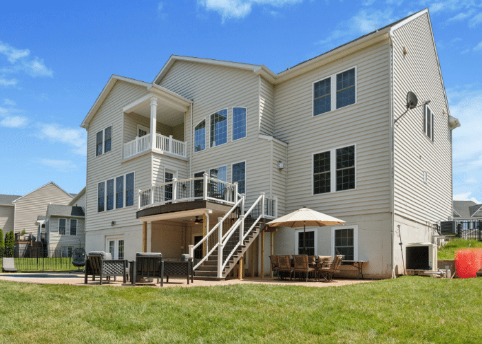 A custom-built elevated deck with white vinyl railings and black balusters, featuring a staircase leading to a large patio area with outdoor seating and a dining setup. The deck is attached to a beige three-story home, surrounded by a spacious lawn and landscaped backyard in Warrington, PA.