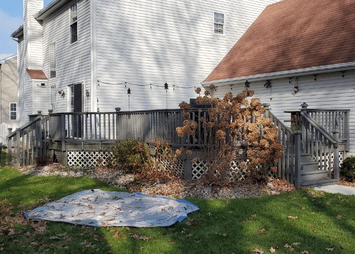 An old wooden deck displays faded brown railings and lattice skirting alongside visible wear and decay. Below the railing hangs a patriotic flag alongside a plastic slide and toy car positioned on the grass.